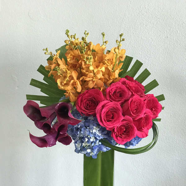 Bright mixed bouquet with pink roses, orange orchids, purple calla lilies, and blue hydrangeas in a tall vase