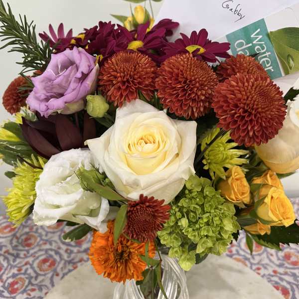 Mixed bouquet of orange mums, white roses, purple lisianthus, and green hydrangea in a glass vase with a card.