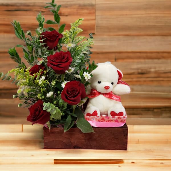 Red roses arranged beside a white teddy bear with a red ribbon