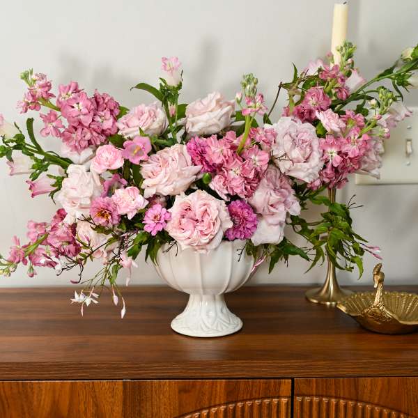 Pink floral arrangement in a white pedestal vase on a wooden cabinet