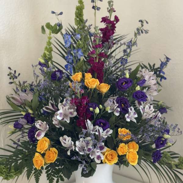 Tall mixed floral arrangement in a white pedestal vase with yellow, purple, and white blooms