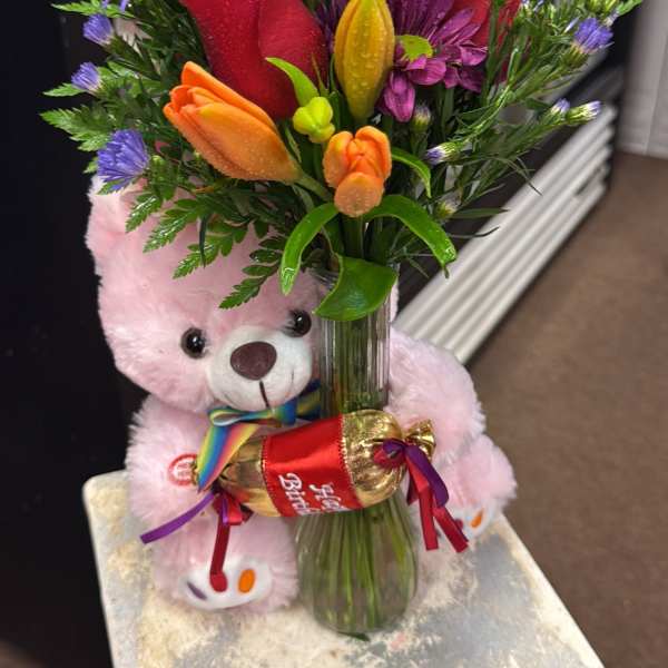 Mixed bouquet in a glass vase with a pink teddy bear and ribboned gift.
