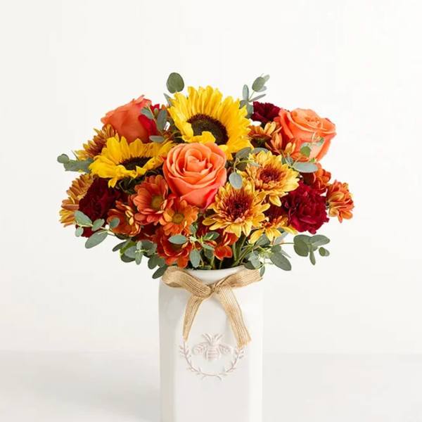 Bouquet of sunflowers, roses, and mums in a white vase with a ribbon