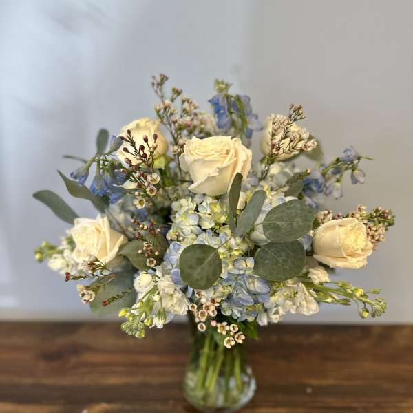 Arrangement of cream roses, blue hydrangeas, and small white flowers in a clear glass vase