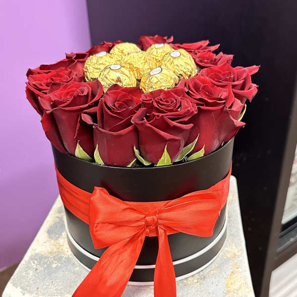 Red roses arranged in a black round box with gold-wrapped chocolates and a red ribbon.