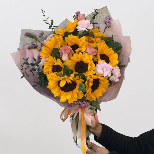 Handheld bouquet of sunflowers, pink roses, and carnations wrapped in pastel paper