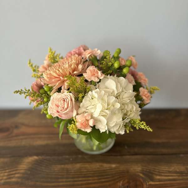 Low arrangement of white hydrangeas and peach roses, carnations, and mums in a clear glass vase