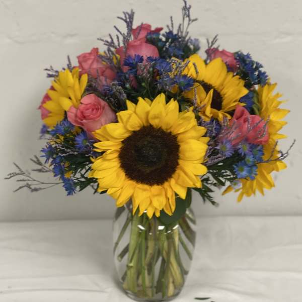 Bouquet of sunflowers, pink roses, and blue filler flowers in a glass vase
