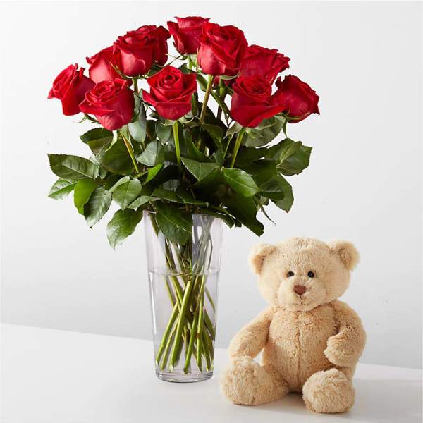 Red roses in a clear glass vase beside a beige teddy bear