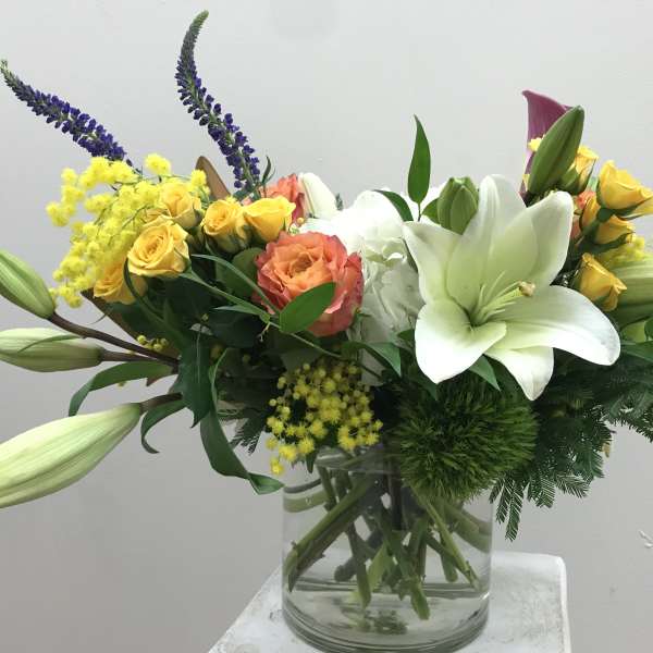 Mixed bouquet with white lilies, yellow roses, and purple spikes in a glass vase