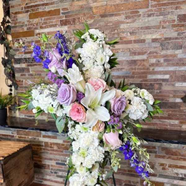 Cross-shaped floral tribute with white lilies, roses, and purple flowers on an easel