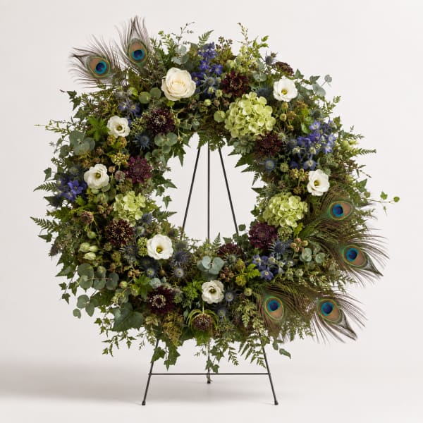 Large floral wreath with white roses, blue flowers, and peacock feathers on a stand