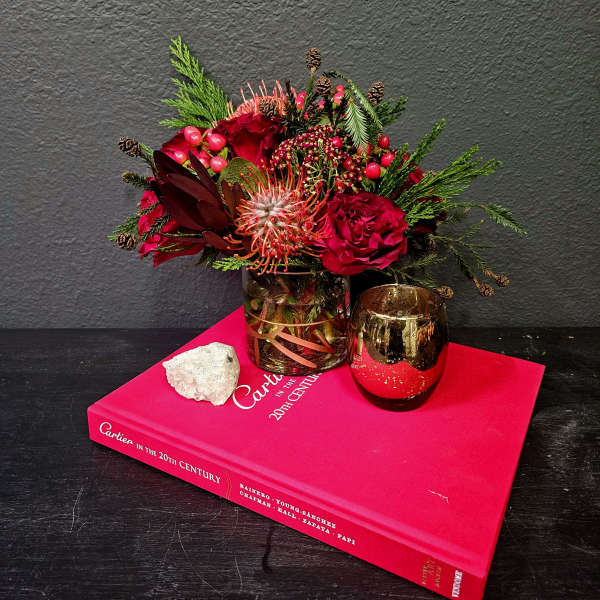 Red floral arrangement in a glass vase beside a gold votive on a pink book