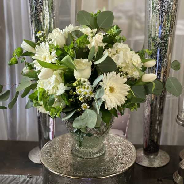 White floral arrangement in a clear glass vase with tall silver candle holders behind it