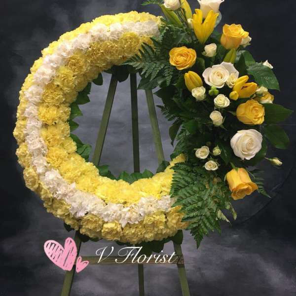 Crescent standing wreath of yellow and white carnations with yellow lilies and roses on a wooden easel.