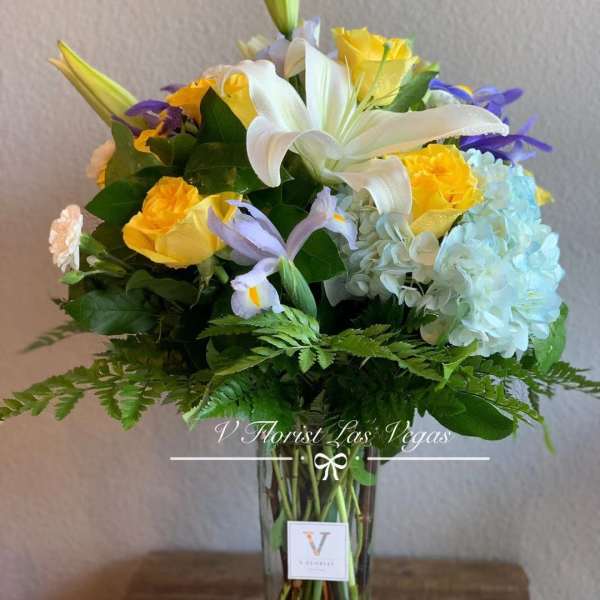 Tall arrangement of white lilies, yellow roses, blue hydrangeas, and purple iris in a clear glass vase
