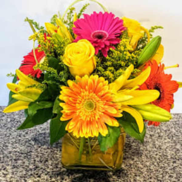 Bright mixed bouquet with gerbera daisies, roses, and lilies in a square vase