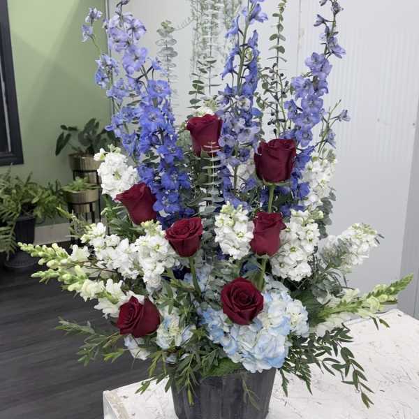 Tall floral arrangement with red roses, blue delphinium, and white blooms in a black container