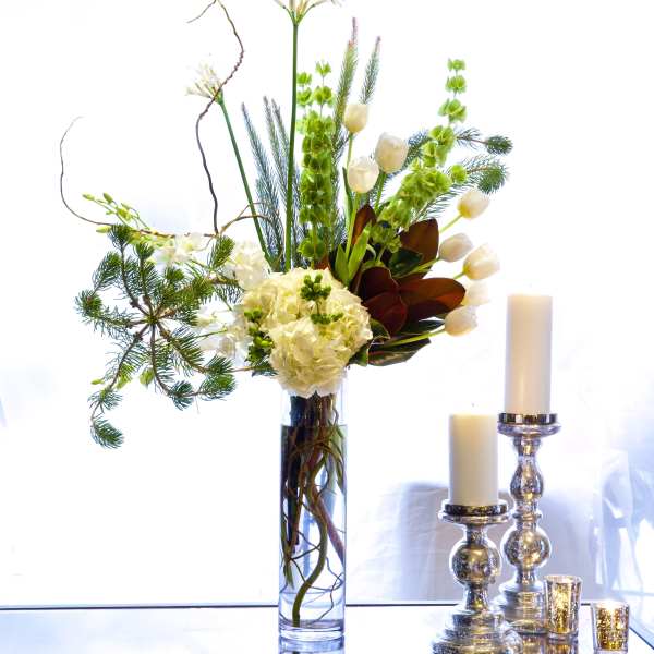 Tall white floral arrangement in a glass vase beside silver candle holders