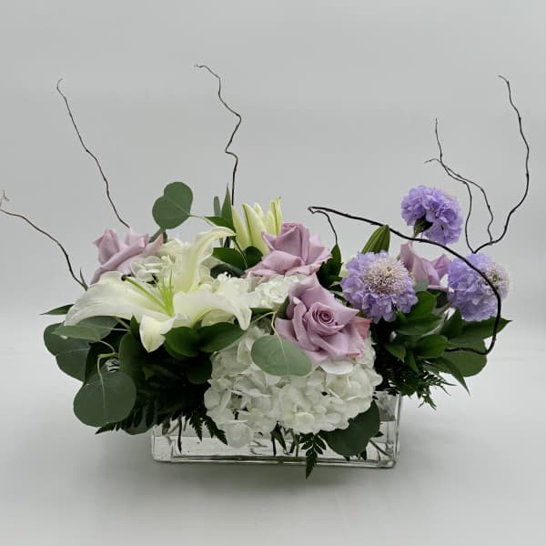 Lavender roses and white lilies arranged with purple blooms in a clear rectangular vase.