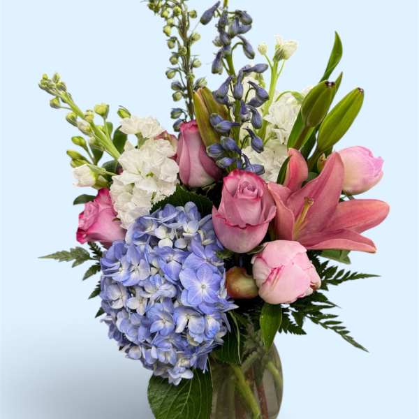 Mixed bouquet of pink roses, blue hydrangea, white blooms, and lilies in a glass vase