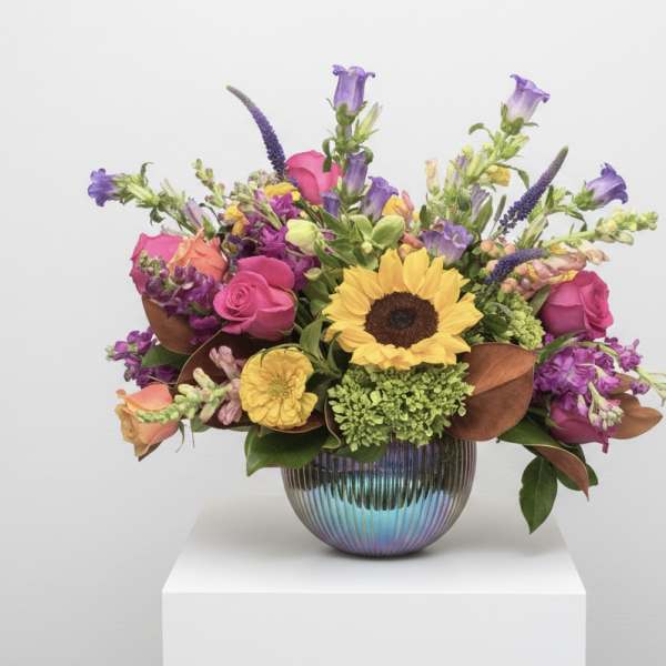 Mixed bouquet with a sunflower, pink roses, and purple blooms in a ribbed iridescent vase