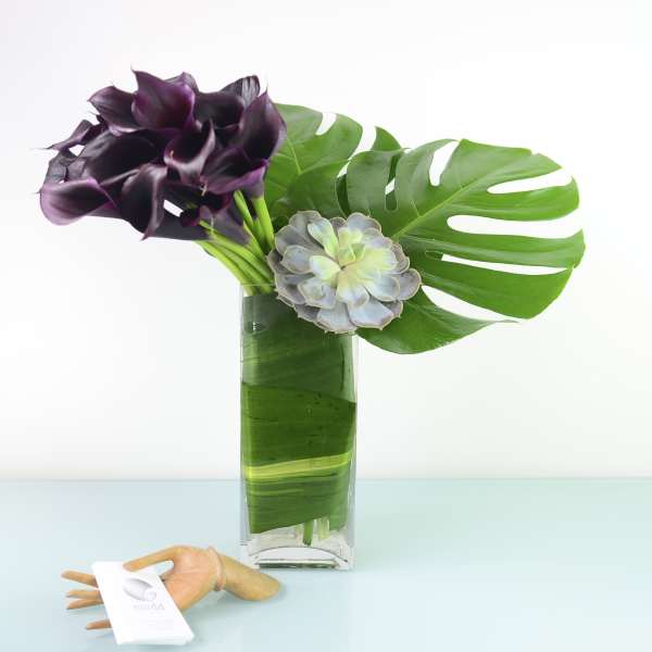 Purple calla lilies and a succulent in a glass vase with large tropical leaves