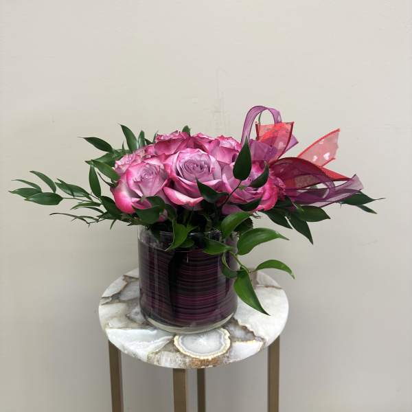 Low arrangement of bright pink roses in a glass cylinder vase with pink ribbon accents.