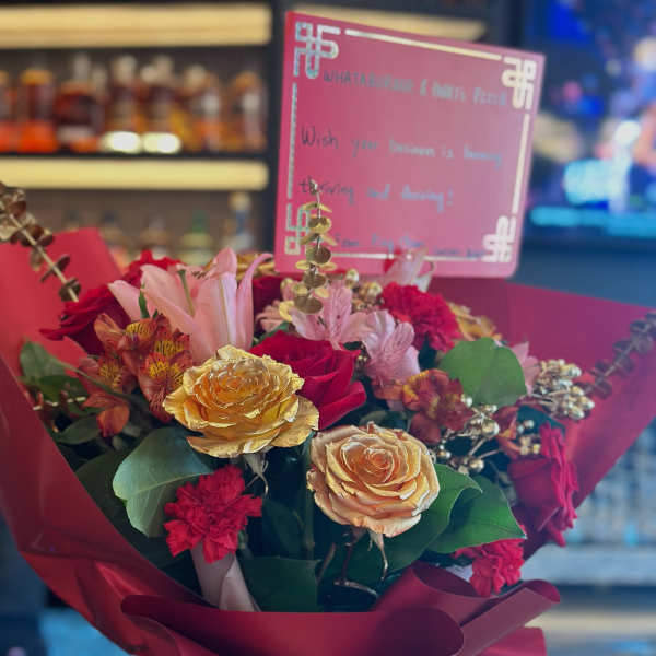Red and gold rose bouquet with pink lilies and mixed blooms wrapped in deep red paper with a message card.