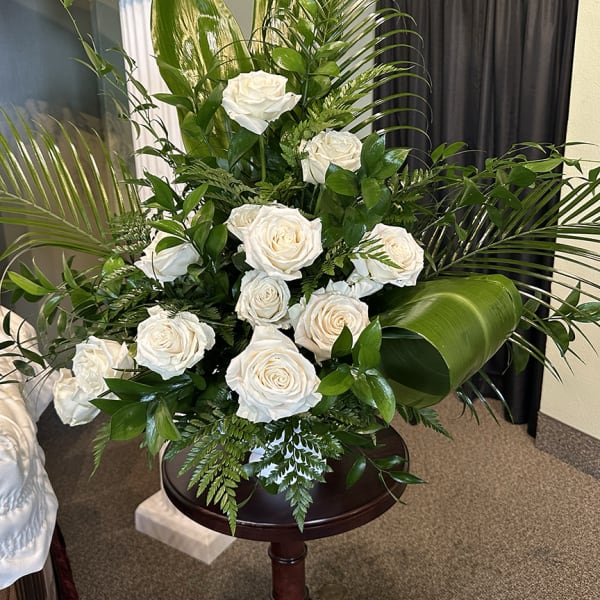 Tall arrangement of white roses with large green tropical leaves on a wooden pedestal.