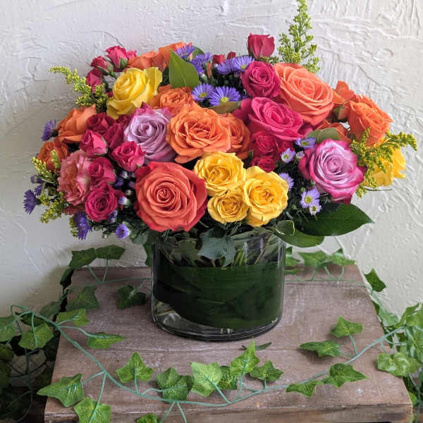 Bouquet of orange, yellow, pink, and lavender roses with purple asters in a clear cylinder vase.