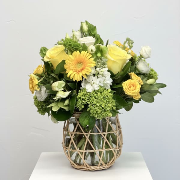 Yellow and white flower arrangement in a glass vase with wicker wrap