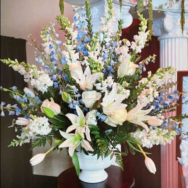 Tall arrangement of white lilies, roses, and blue delphinium in a white pedestal vase