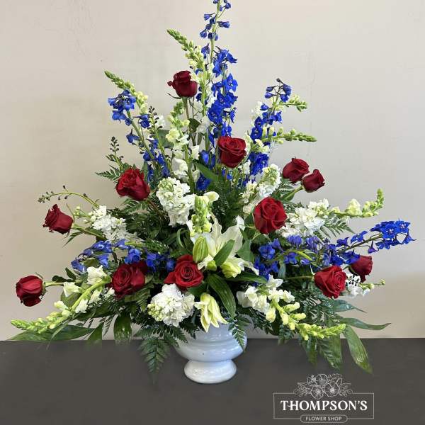 Tall red, white, and blue arrangement with roses, lilies, and delphinium in a white urn vase