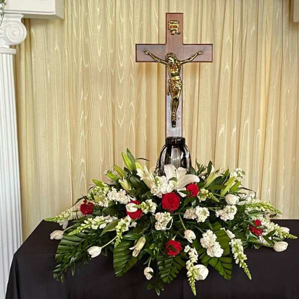 Sympathy floral arrangement with red roses, white lilies, and a crucifix backdrop