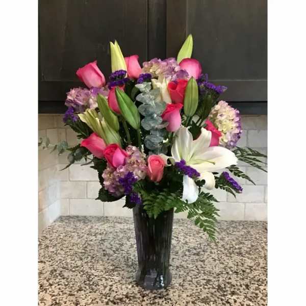 Tall vase arrangement of pink roses, white lilies, and purple flowers in a dark glass vase