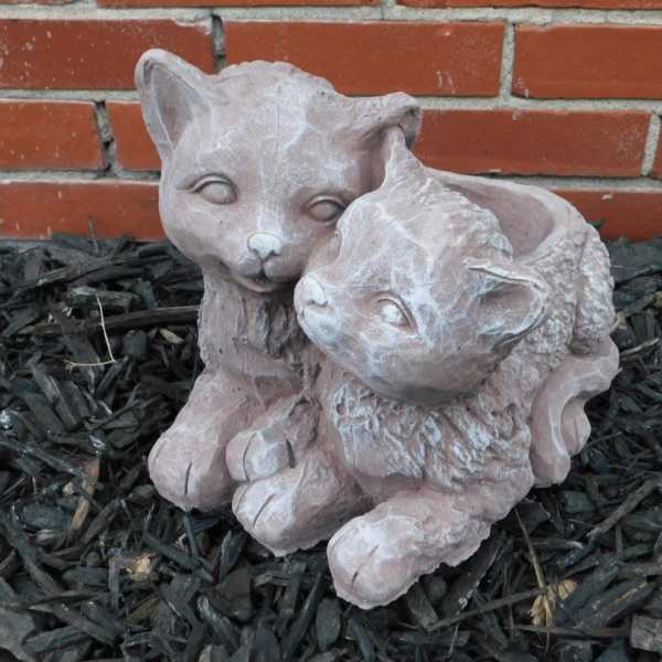 Two small cat figurines sitting together on mulch in front of a brick wall.