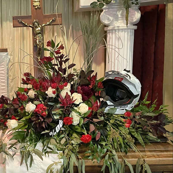 Red and white floral casket spray with roses and carnations displayed beside a white motocross helmet.