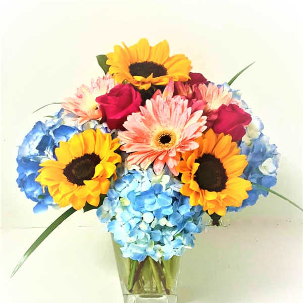 Bouquet of sunflowers, pink gerberas, roses, and blue hydrangeas in a clear vase