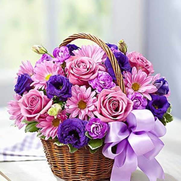 Pink and purple roses, daisies, and blooms arranged in a wicker basket with a lavender bow