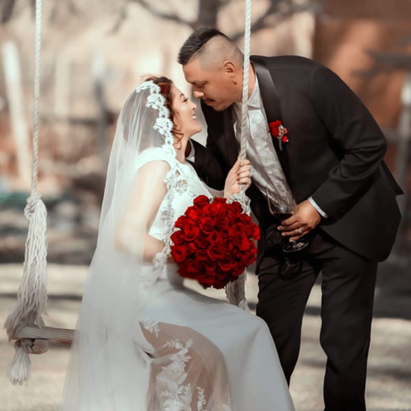 Bride holding a red rose bouquet while leaning toward groom on a swing