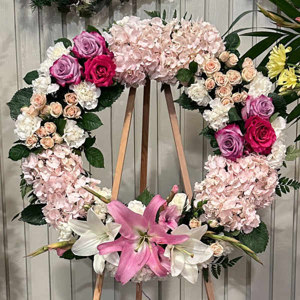 Heart-shaped floral wreath with pink hydrangeas, roses, and lilies on a wooden stand