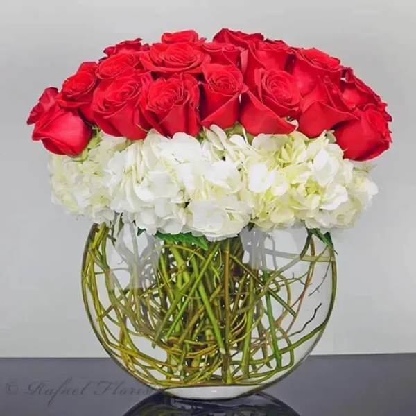 Round arrangement of red roses over white hydrangeas in a clear glass bowl vase with exposed stems