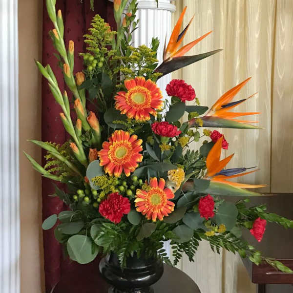 Tall arrangement of orange gerberas, red carnations, and birds of paradise in a black urn vase