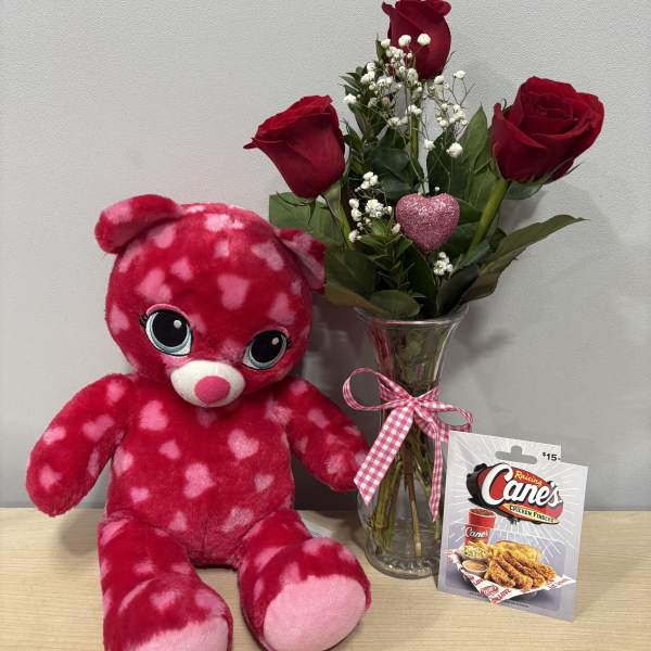 Red roses in a glass vase beside a pink heart-pattern teddy bear and gift card