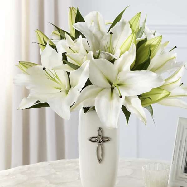 White lilies arranged in a tall white vase with a cross emblem