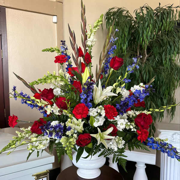 Tall red, white, and blue funeral arrangement with roses, lilies, carnations, and delphinium in a white urn.