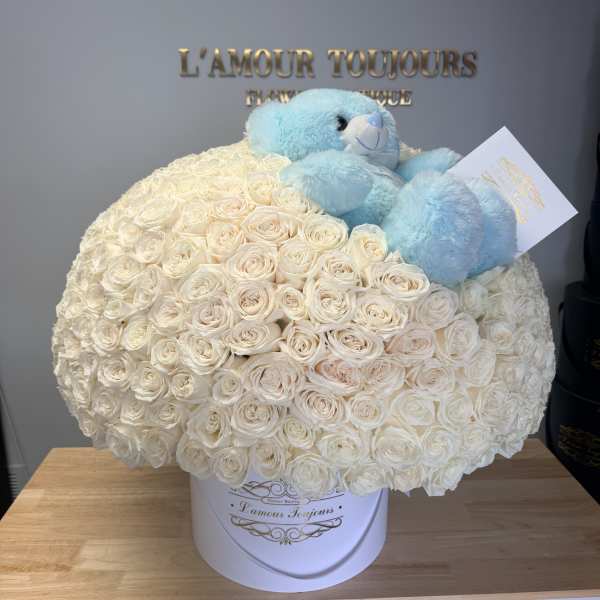 White rose arrangement in a round box with a blue teddy bear on top