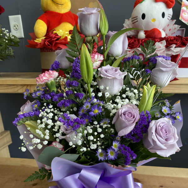 Lavender roses and lilies arranged with purple filler flowers and a satin ribbon bow.