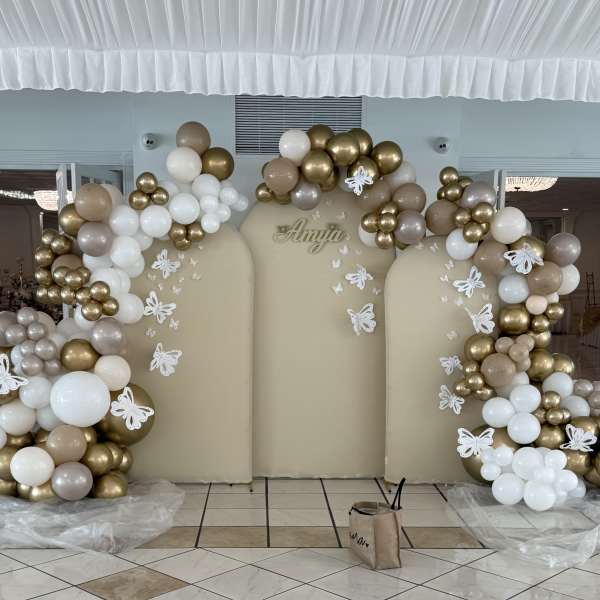 Gold, white, and beige balloon arch around a backdrop with butterfly decorations